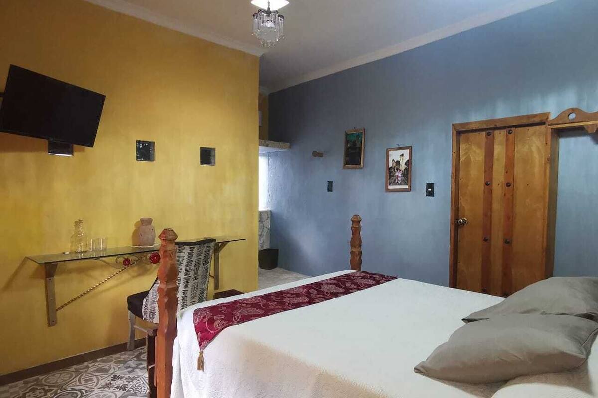 A bedroom with a bed, a wooden wardrobe, a television on the wall, and a small table with a chair.