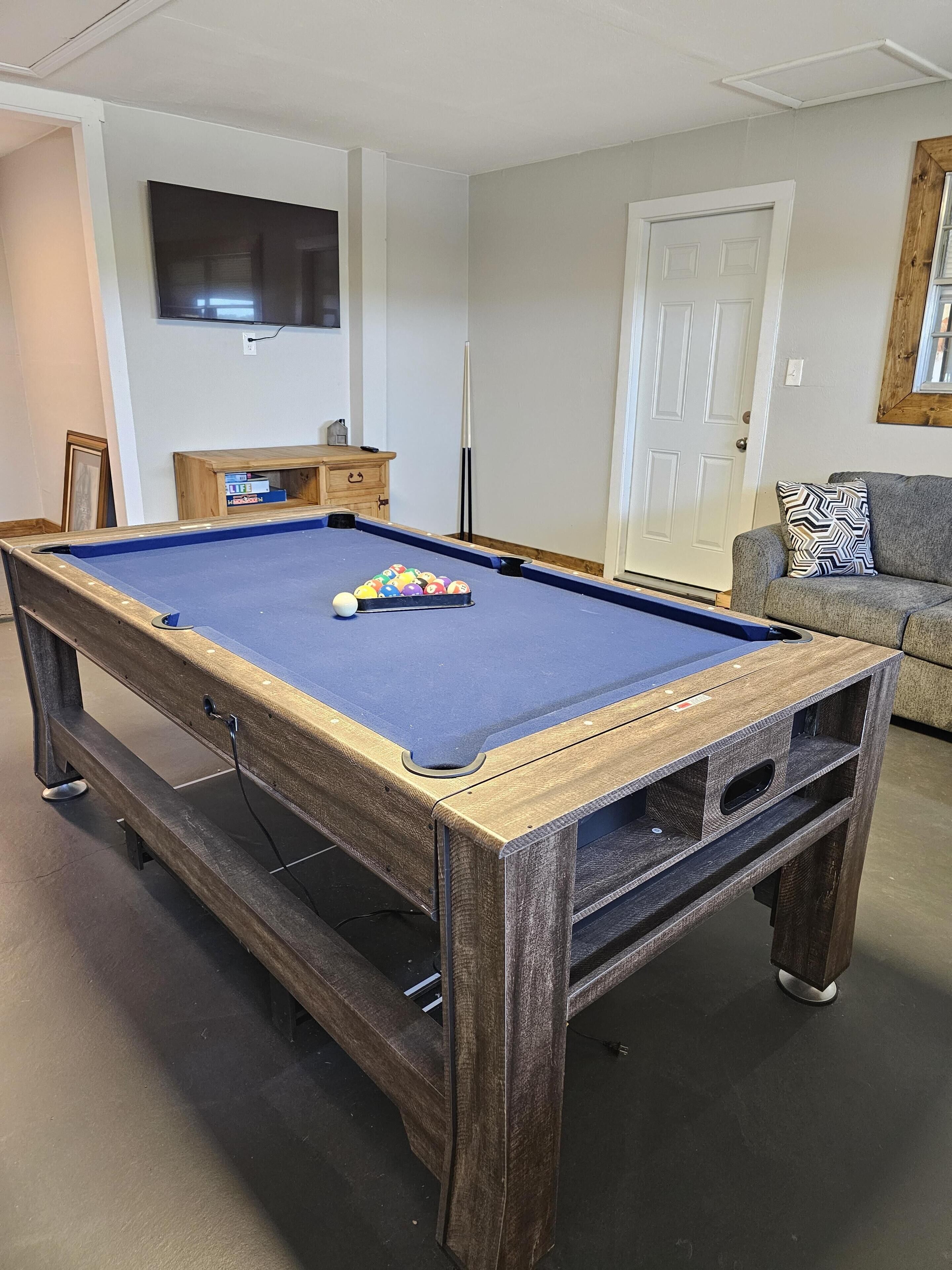 Un biliardo con struttura in legno e superficie rivestita di feltro blu, una televisione fissata al muro, un mobiletto in legno e un divano grigio con un cuscino decorato.