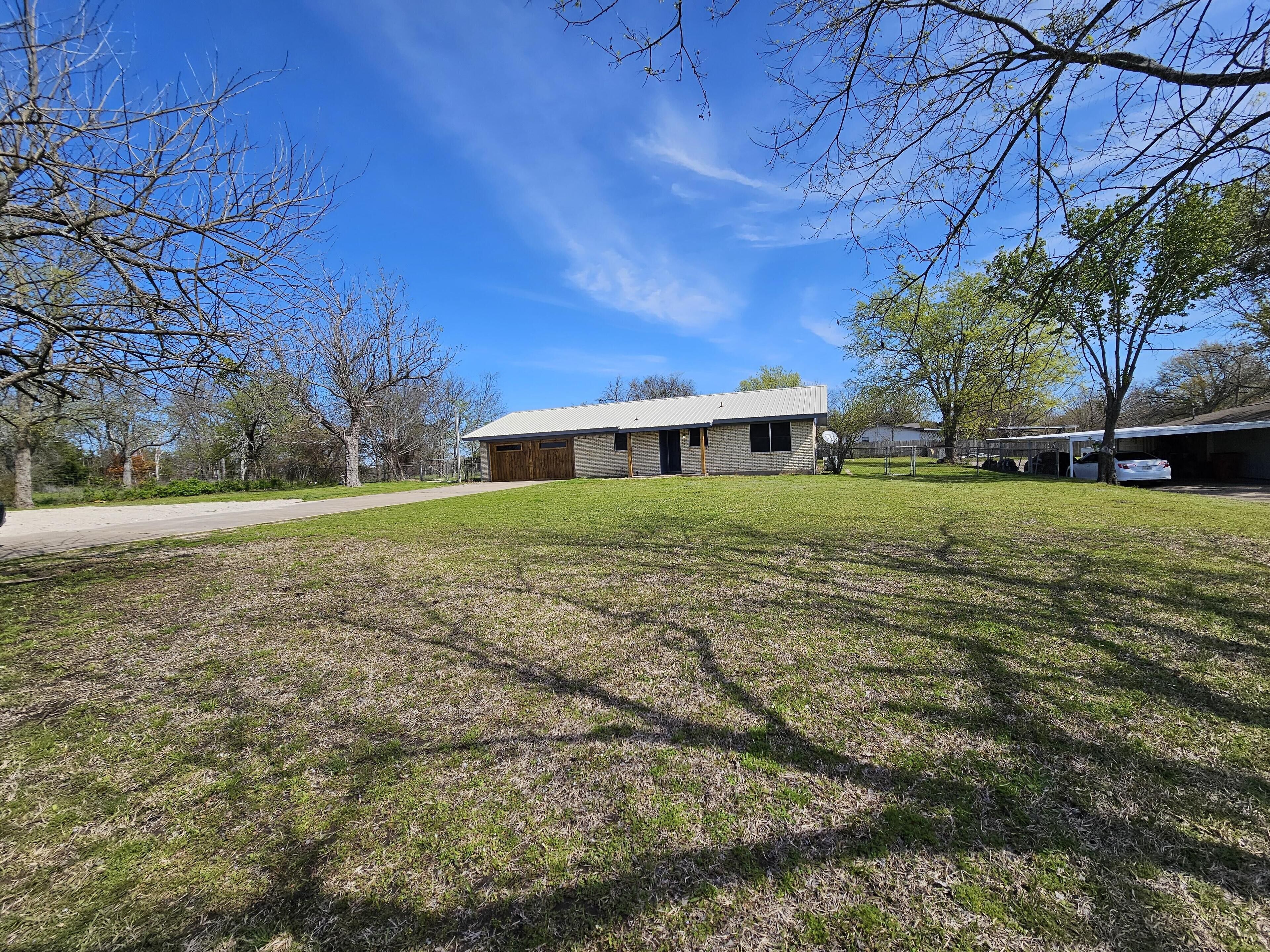 A single-story house with a brick exterior and a metal roof, surrounded by a grassy yard and trees.