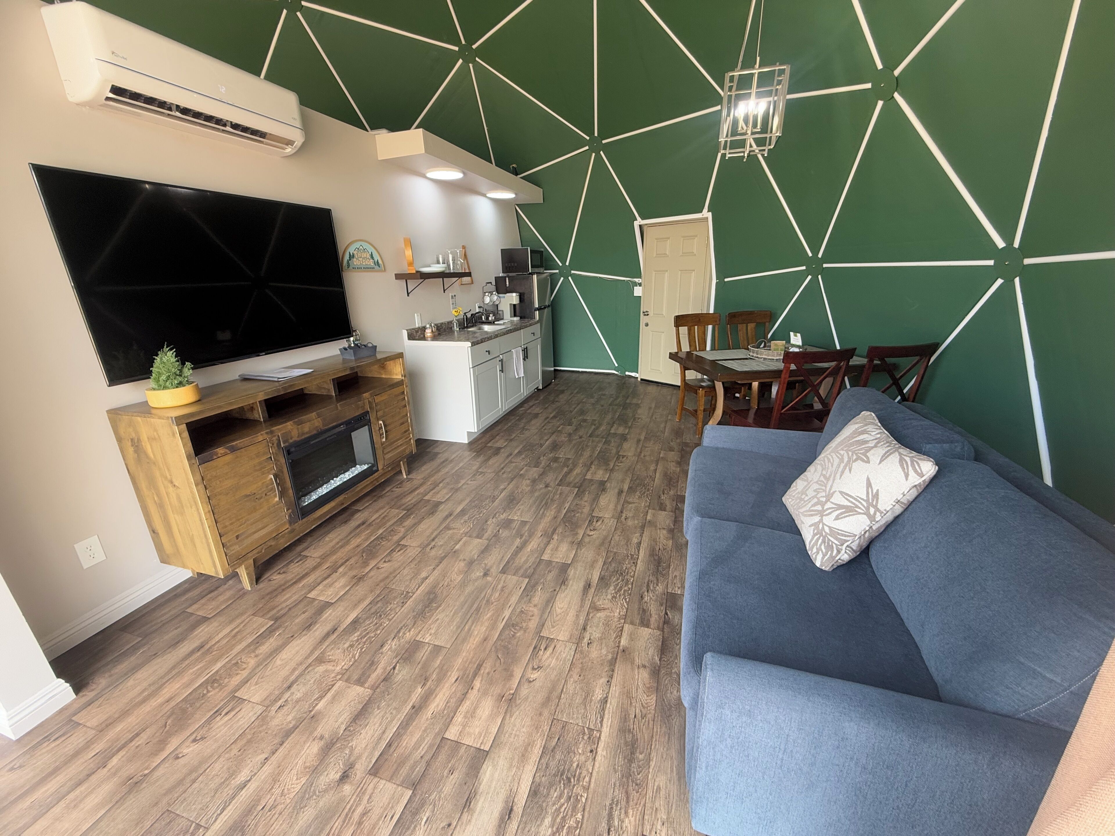 A modern living room with a flat-screen TV, wooden entertainment unit, blue sofa, dining table with chairs, kitchen area, and a geometric-patterned wall.