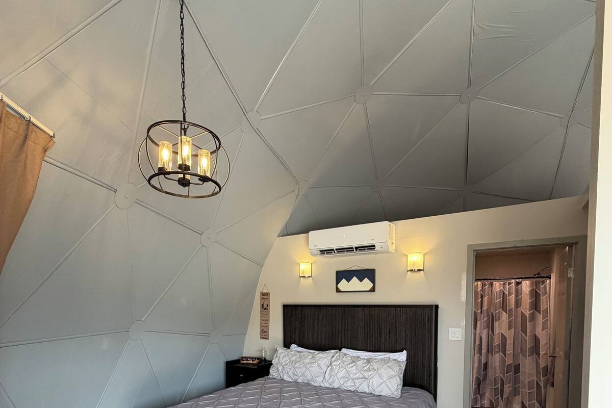 Une chambre avec un plafond en pente, un lit recouvert d’une courtepointe blanche, un luminaire suspendu et un climatiseur fixé au mur.
