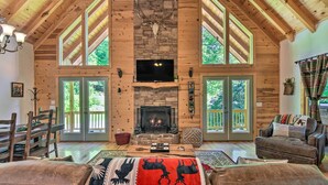 A spacious living room with a stone fireplace, wooden beams, and large windows. - Great for Families! Lavish Hiawassee Cabin (Hiawassee)