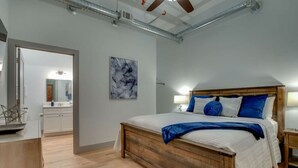 Ein Schlafzimmer mit einem hölzernen Bett, weißer Bettwäsche und blauen Kissen. Im Hintergrund ist ein Badezimmer zu sehen.
