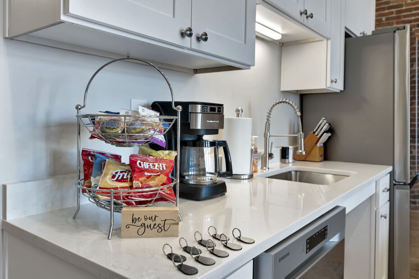 A kitchen with a coffee maker, a snack basket, and a 'be our guest' sign.