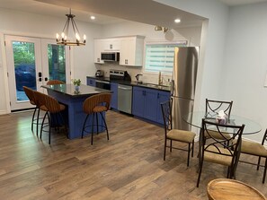 Uma cozinha moderna com uma ilha central, armários azuis, eletrodomésticos de aço inoxidável e um espaço de refeições com mesa e cadeiras de vidro.
