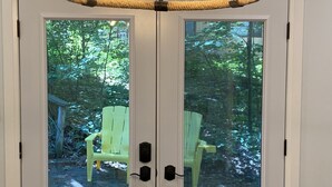 A room with a white door featuring two glass panels, a chandelier with exposed bulbs, and a view of a green outdoor area with a yellow chair. - Cozy Sandy Pines Cottage Suite, right on Lake Huron in a Carolinian Forest,  (Grand Bend)
