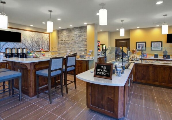 A breakfast bar with a sign stating 'FREE BREAKFAST INCLUDED,' bar stools, a stone wall, and a television mounted on the wall.