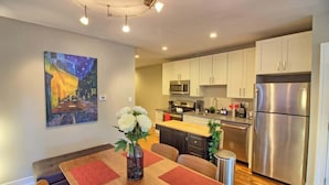 A modern kitchen with stainless steel appliances, a dining area with a wooden table, and a painting on the wall. - Stunning Minimalist Gem | Fenced Backyard + W/D (Saint Louis)