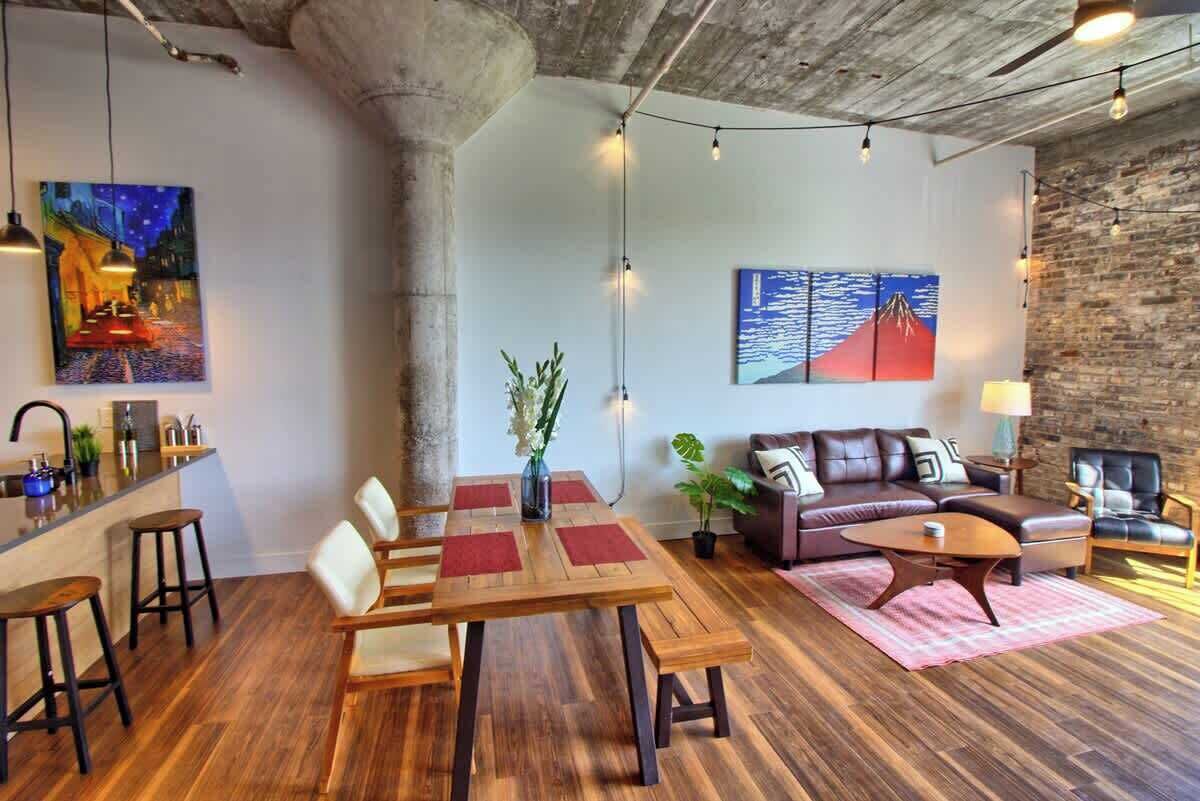 A modern living room with a wooden dining table, a sofa, and a coffee table. There are paintings on the walls, a plant, and a bar area with stools.