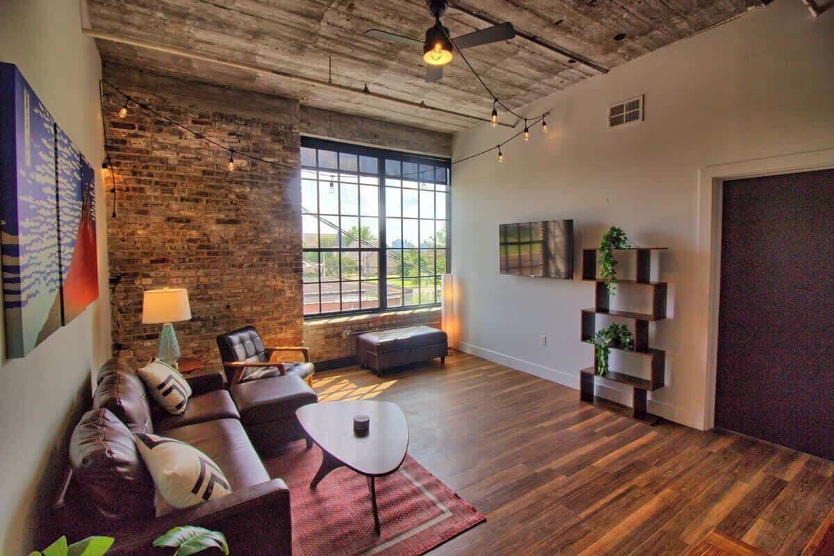 A spacious living room with a brick wall, large windows, a sofa, a coffee table, and a bookshelf.