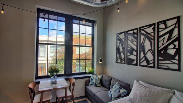 A room with a large window, a sofa, a table with chairs, and abstract wall art. - Chic Ultramodern Studio Loft w/ Designer Features (Saint Louis)