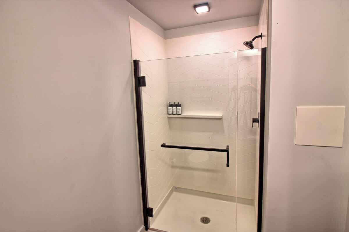 A glass-enclosed shower stall with a black door handle, a glass shower door, and a shelf with toiletries.