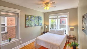 A bedroom with a bed, a ceiling fan, two windows, and a brick wall. - Irises at TG Park | Fenced Backyard+W/D+Storage (Saint Louis)