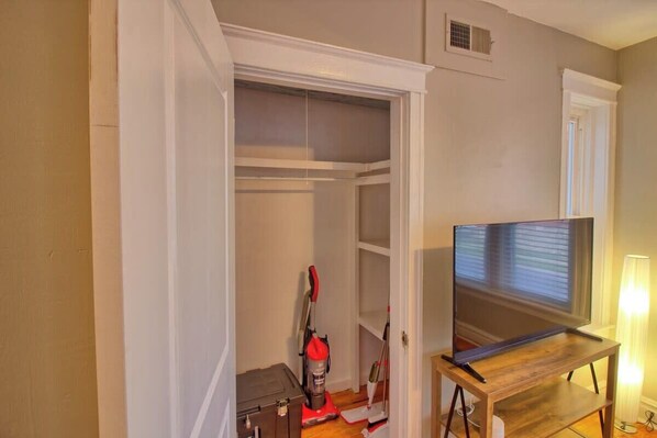 A room with a television on a wooden stand, a closet with white doors, and cleaning supplies.