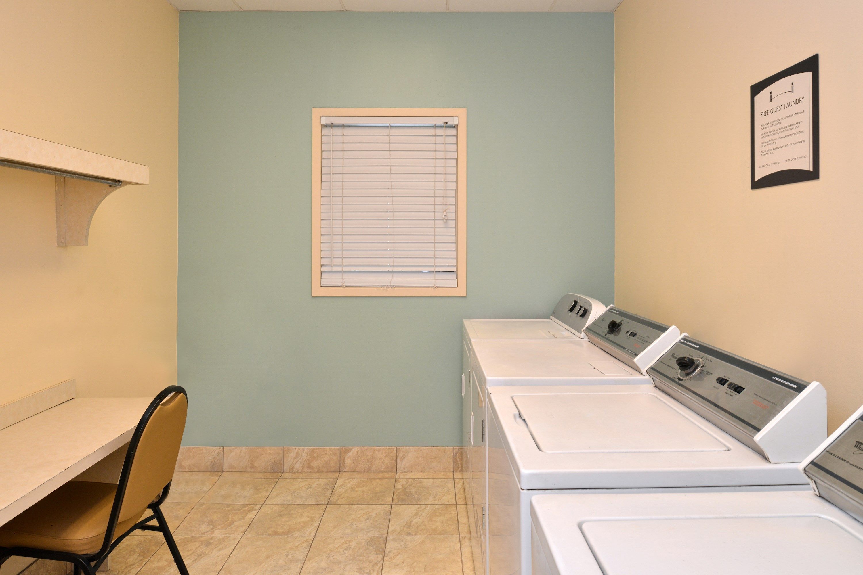 A laundry room with a chair, a window with blinds, and a sign indicating free guest laundry.