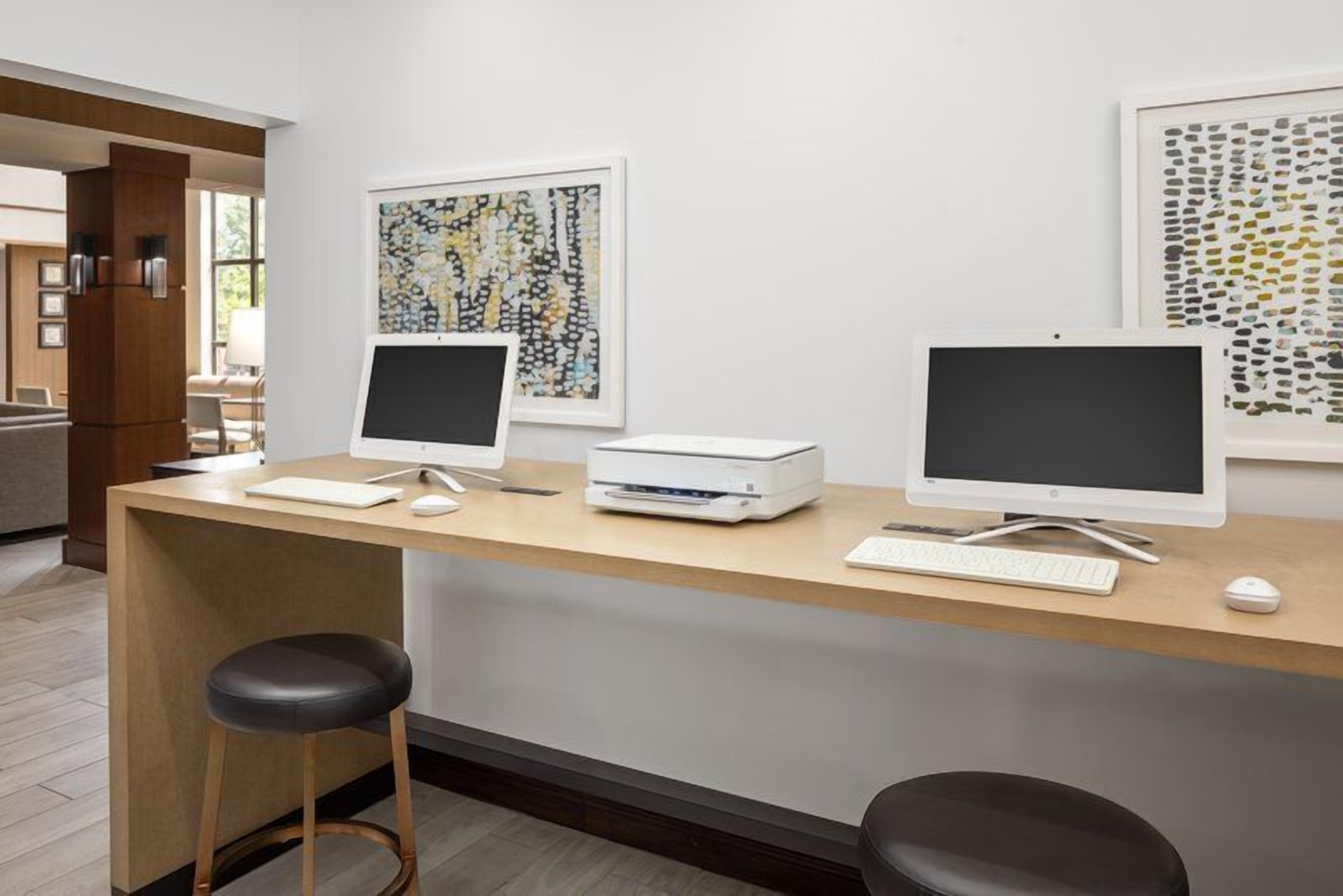 A reception desk with two monitors, a printer, a keyboard, and a mouse. There are two stools and framed abstract art on the wall.