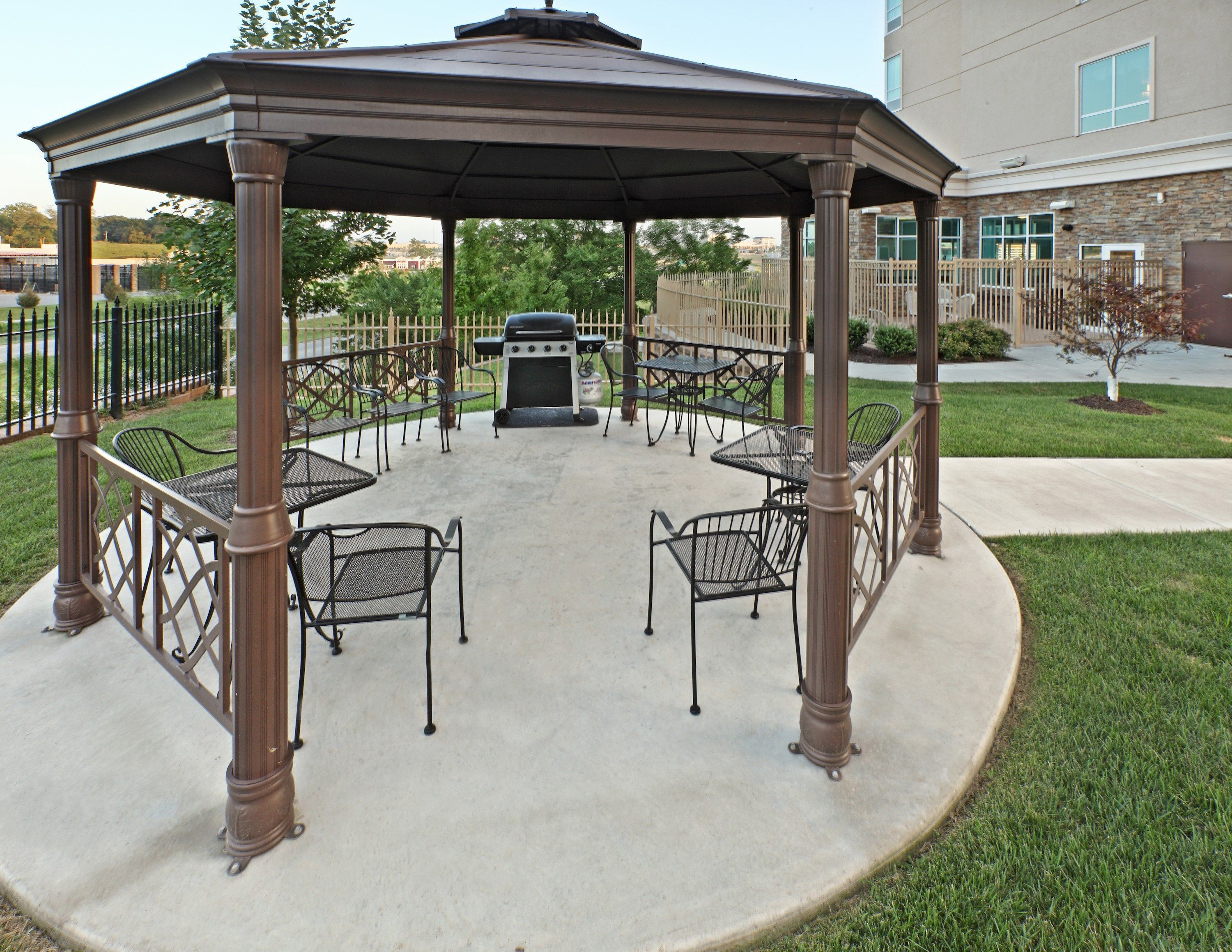 En pergola med fyra metallstolar och en grill, omgiven av en betongterrass och en stenbyggnad i bakgrunden.