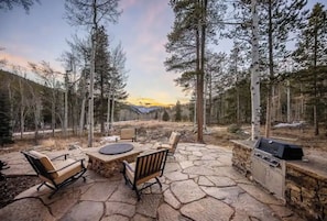 Une terrasse extérieure avec un sol en pierre, une table et des chaises, entourée d’arbres et offrant une vue sur une chaîne de montagnes.