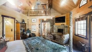 Acogedora habitación tipo cabaña de madera con cama en altillo, chimenea y un ventanal grande.