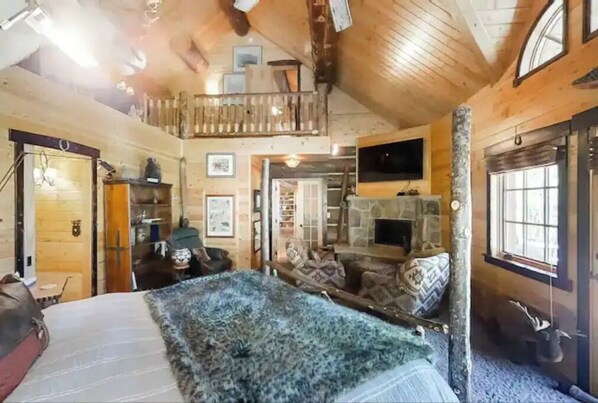 Une chambre chaleureuse dans un chalet en rondins, avec un lit en mezzanine, une cheminée et une grande fenêtre.