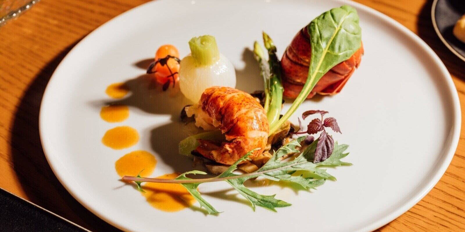 A white plate with a variety of food items including a piece of meat, vegetables, and a small orange sphere, arranged on a wooden table.