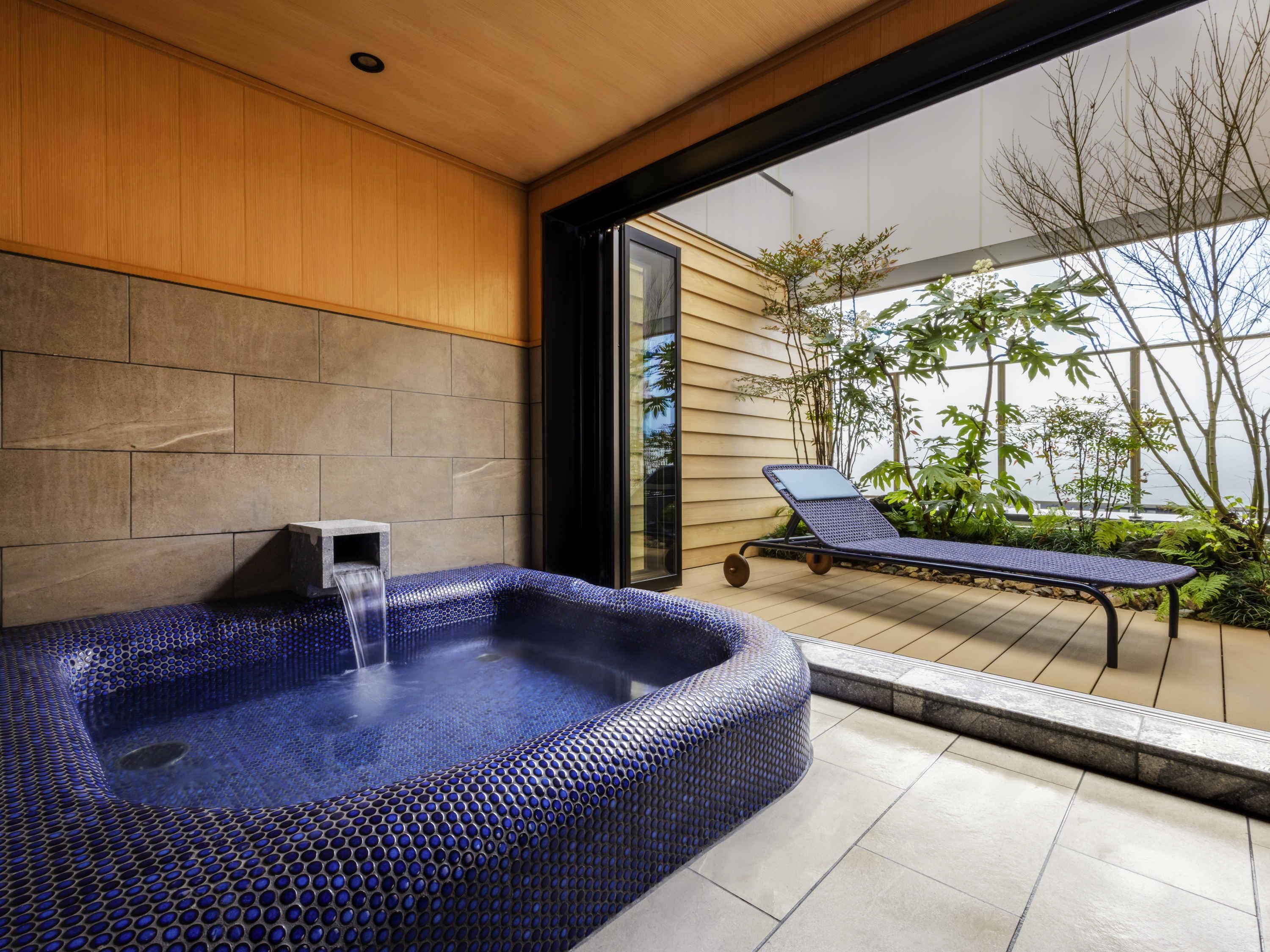 Ein moderner Wellnessbereich mit Whirlpool, Liege und Blick auf einen Garten durch eine Glastür.