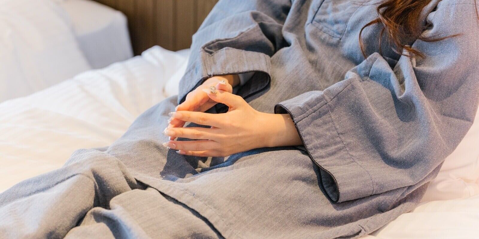 Una persona con una bata gris sentada sobre una cama con ropa blanca.