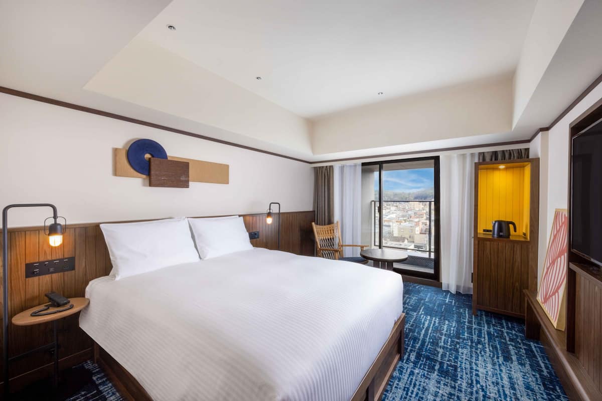 A hotel room with a large bed, a desk with a telephone, a window with a city view, and a small table with a chair.