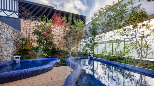 A modern outdoor pool area with two hot tubs, surrounded by a stone wall and lush greenery. - Mercure Hida Takayama (Takayama)