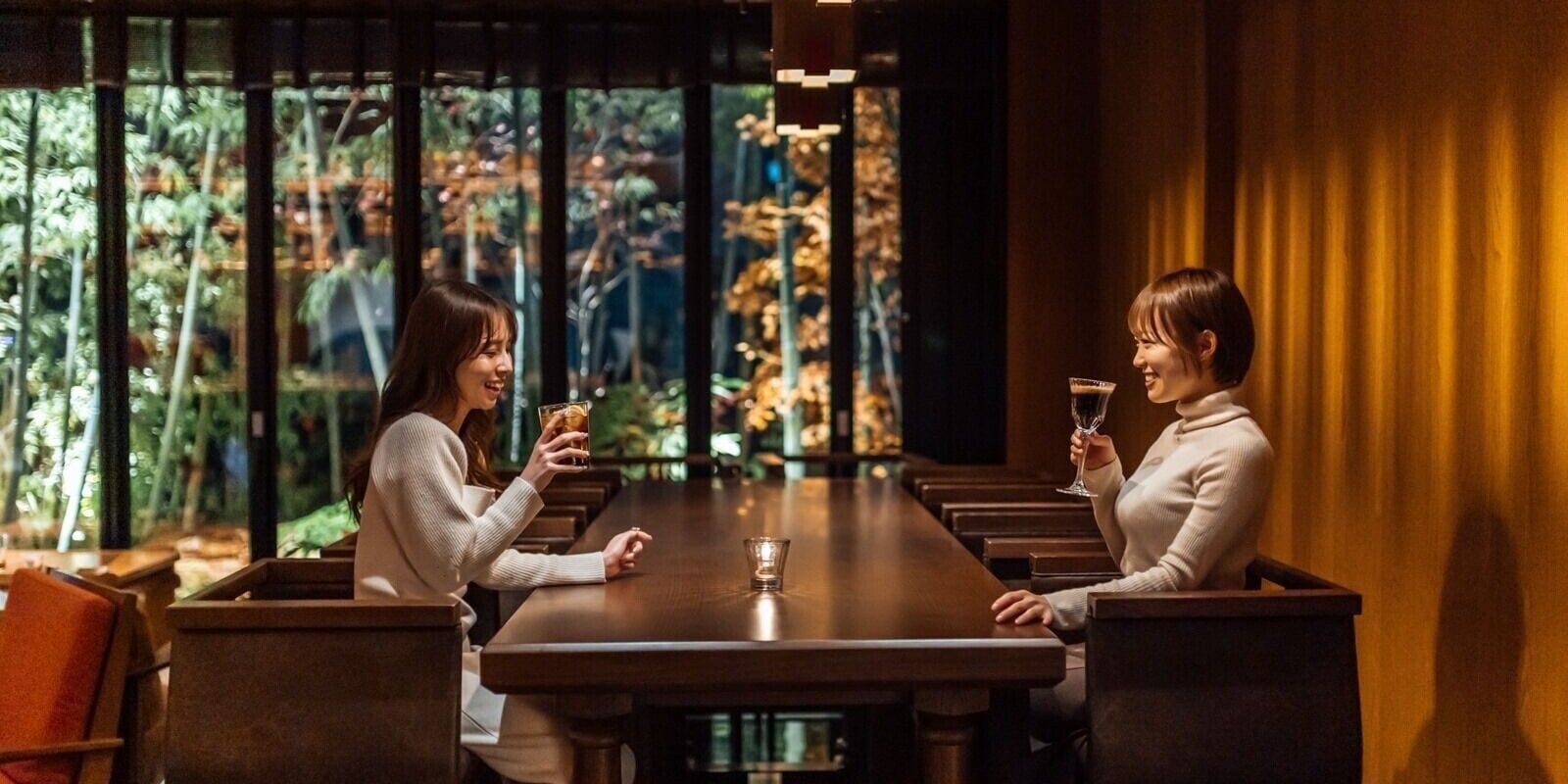 Two people seated at a long wooden table with a glass of drink each, in a room with large windows and a view of greenery outside.