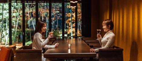 Two people seated at a long wooden table with a glass of drink each, in a room with large windows and a view of greenery outside.