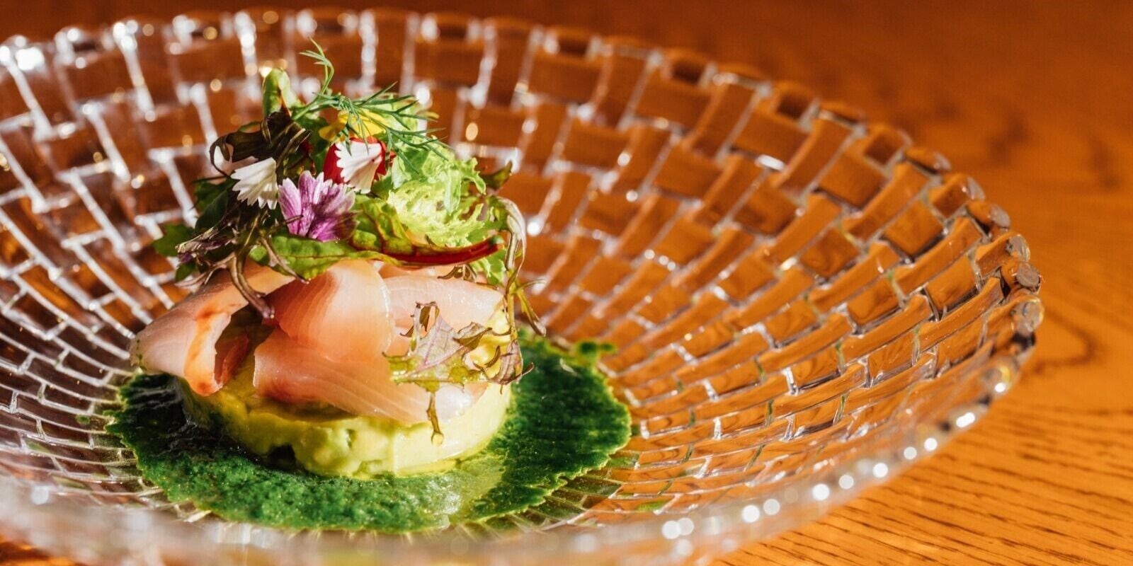 Een glazen schaal met een gelaagde schotel, mogelijk een salade of voorgerecht, op een houten ondergrond.