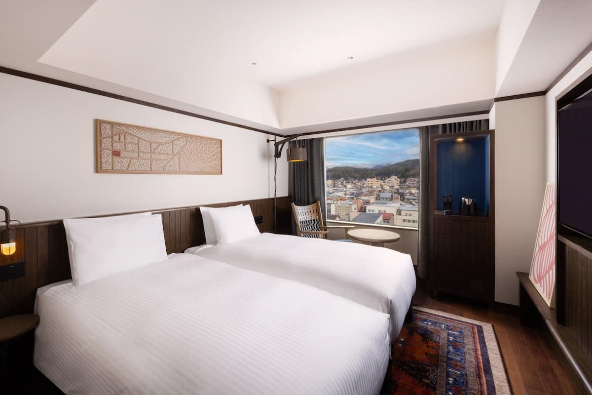 A hotel room with two beds, a large window with a city view, a small round table, and a decorative wall hanging.