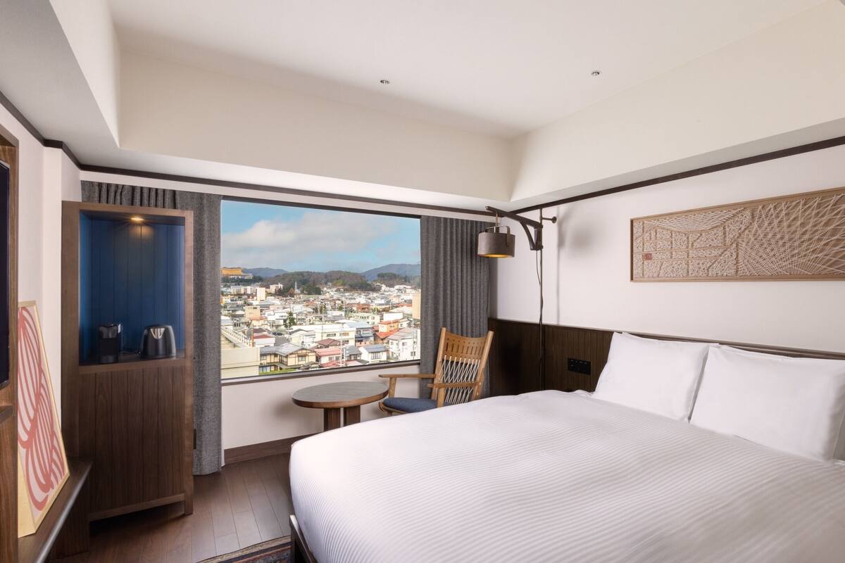 A hotel room with a large bed, a view of a cityscape, a small table, and a chair.