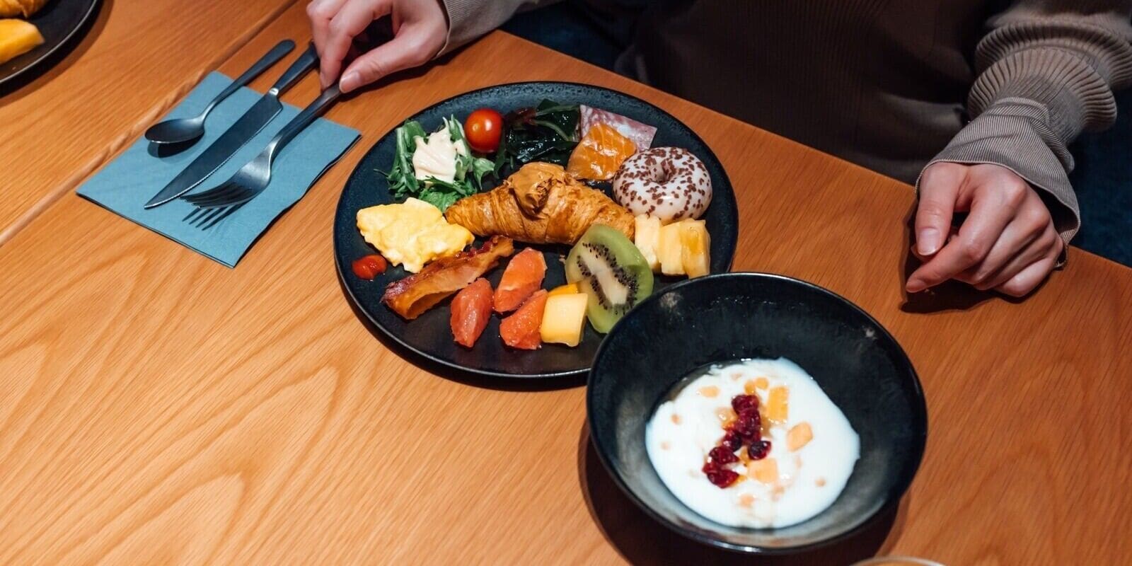 Una mesa de madera con un plato de comida, un bol de yogur y un mantel con cubiertos.