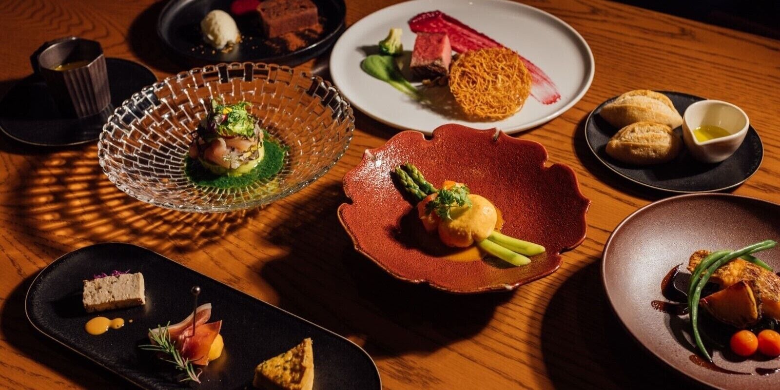 Diversi piatti su un tavolo di legno, tra cui una ciotola di vetro con guarnizione verde, un piatto rosso con asparagi e un piatto nero con un piccolo dessert.