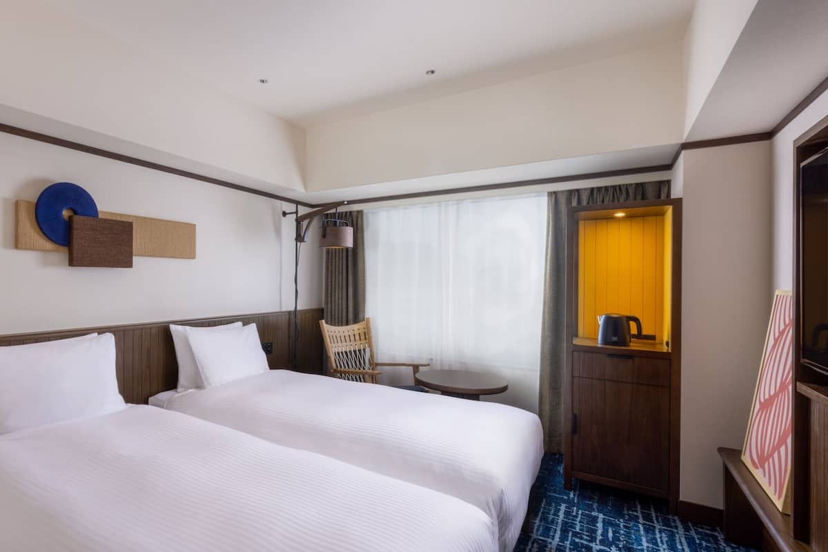A hotel room with two beds, a small table, a chair, a lamp, and a window with curtains.
