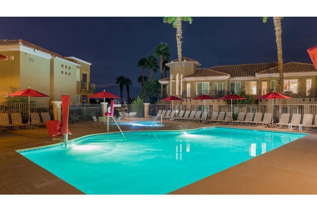A swimming pool with lounge chairs and umbrellas, surrounded by buildings with palm trees.