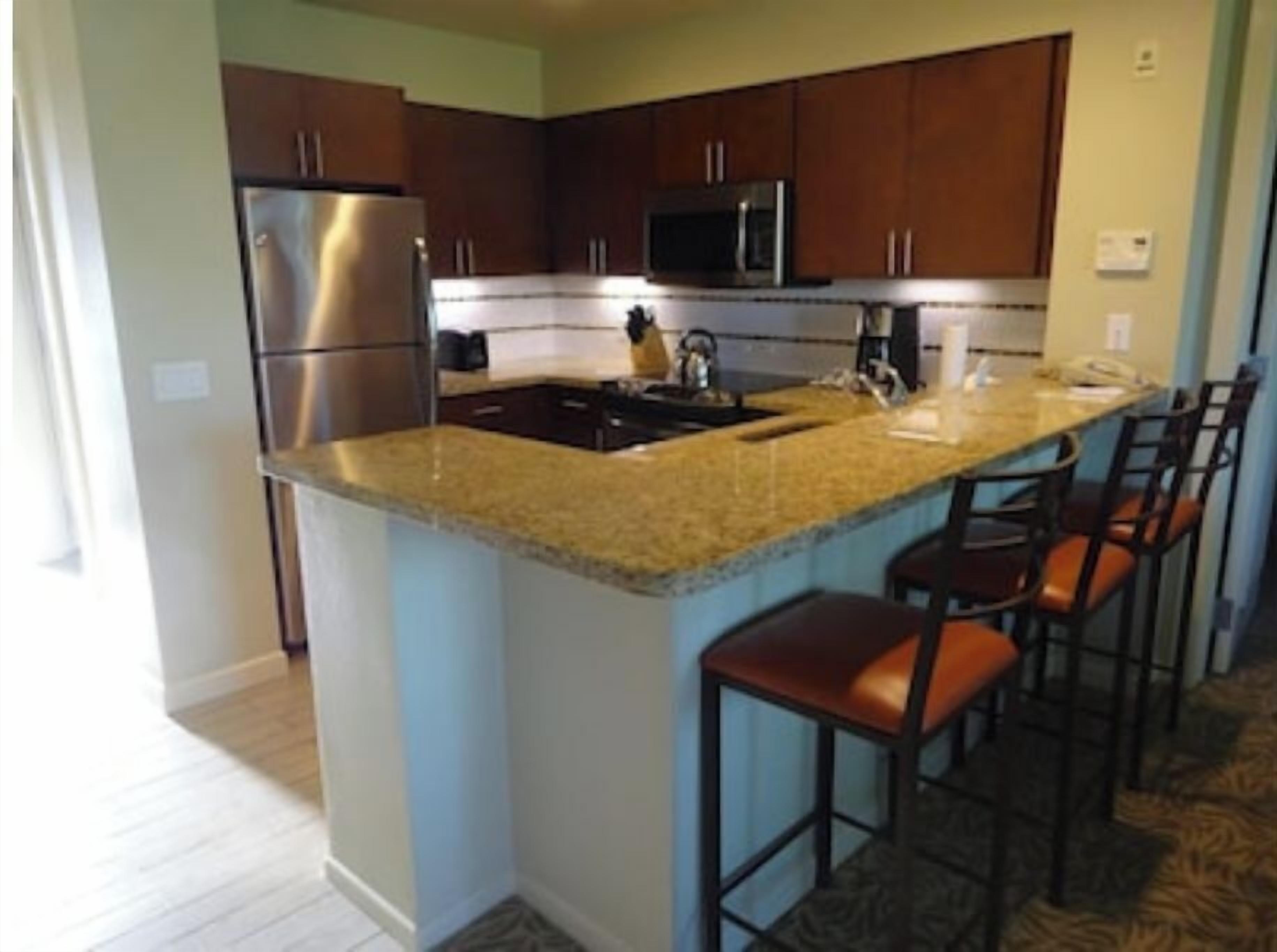 A kitchen with a stainless steel refrigerator, a microwave, a sink, and a granite countertop. There are four bar stools with leather seats.