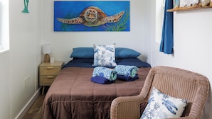 Un dormitorio con cama, mesita de noche, una silla de mimbre, un cuadro de una tortuga marina y un estante con objetos decorativos.
