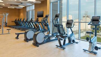 A gym with treadmills, elliptical machines, and stationary bikes, large windows with a city view, and modern lighting fixtures.
