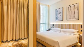 A hotel room with a large bed, a bedside table, a nightstand, and a window with curtains. There are two framed artworks on the wall.