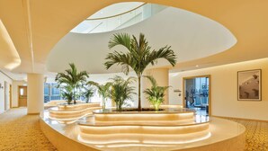 Un vestíbulo moderno con un área de estar circular central, palmeras y un techo alto con tragaluces.