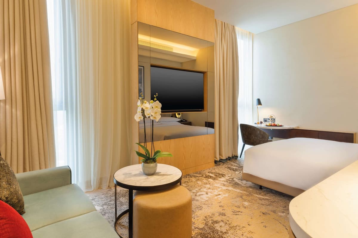 A hotel room with a bed, a desk, a chair, a round table with a plant, and a TV.