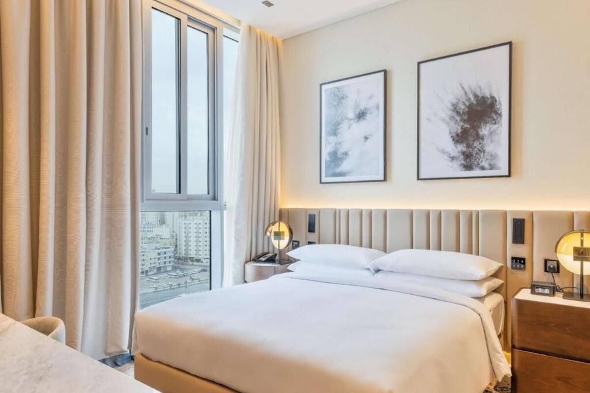 A hotel room with a large bed, two bedside tables with lamps, a view of a cityscape through a window, and two framed abstract artworks on the wall.