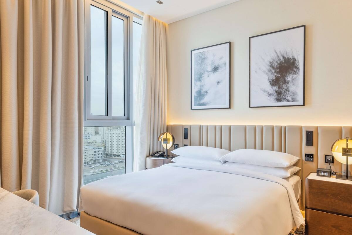 A modern hotel room with a large bed, a bedside table with a lamp, and a view of the city through the window.