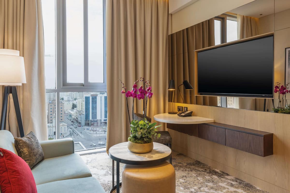 A modern hotel room with a large window, a flat-screen TV, a round table with a potted plant, a sofa, and a bedside table with a lamp.