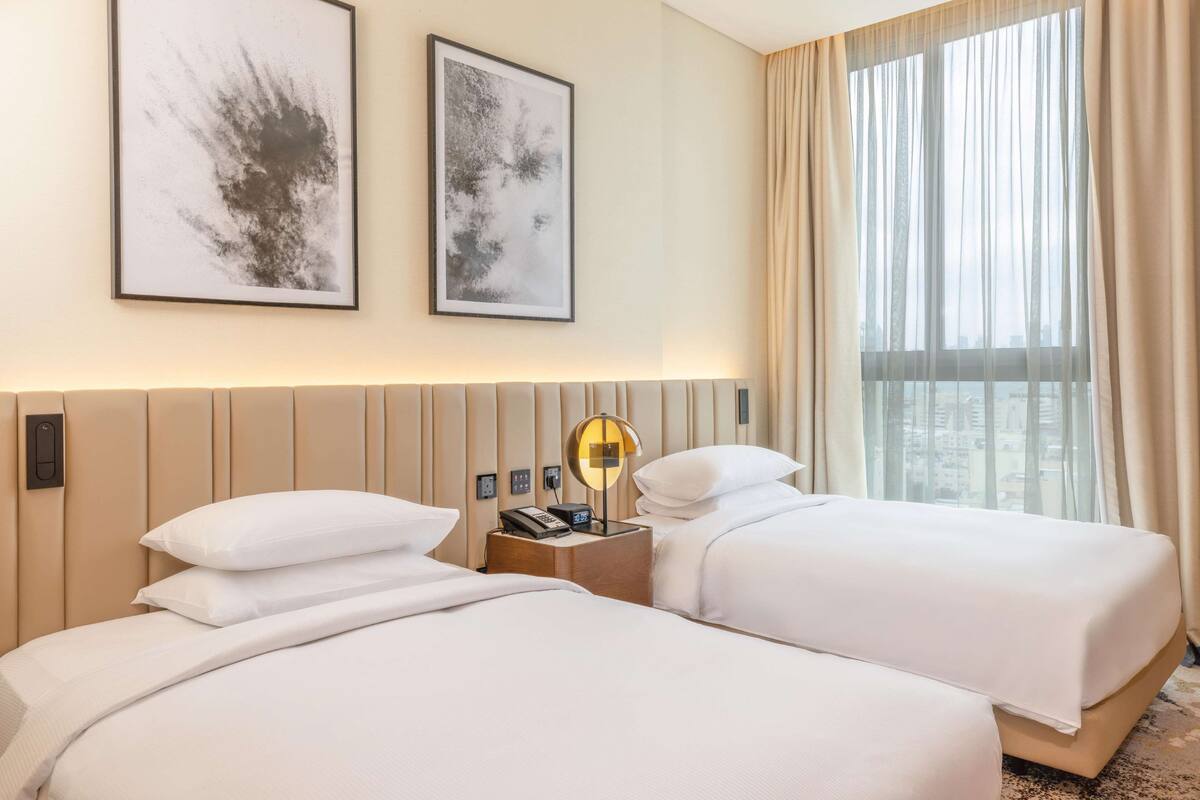 A hotel room with two beds, a nightstand with a lamp, a phone, and a wall-mounted control panel.