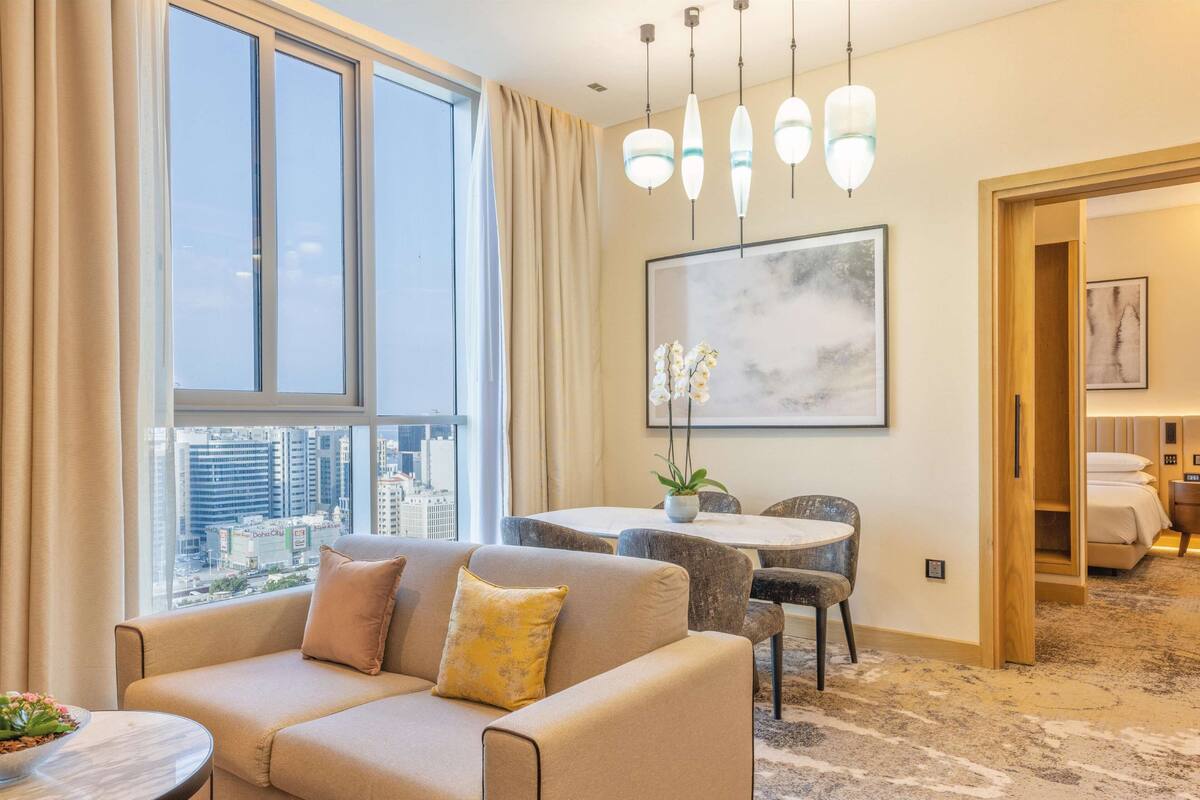 A modern hotel room with a large window, a dining area, and a bed.