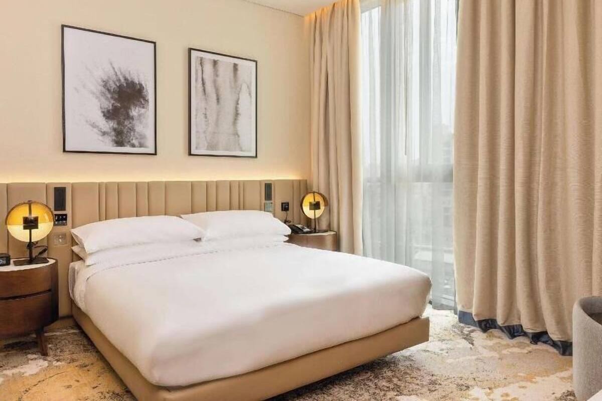 A hotel room with a large bed, two wall-mounted artworks, a nightstand with a lamp, and a view of the city through sheer curtains.